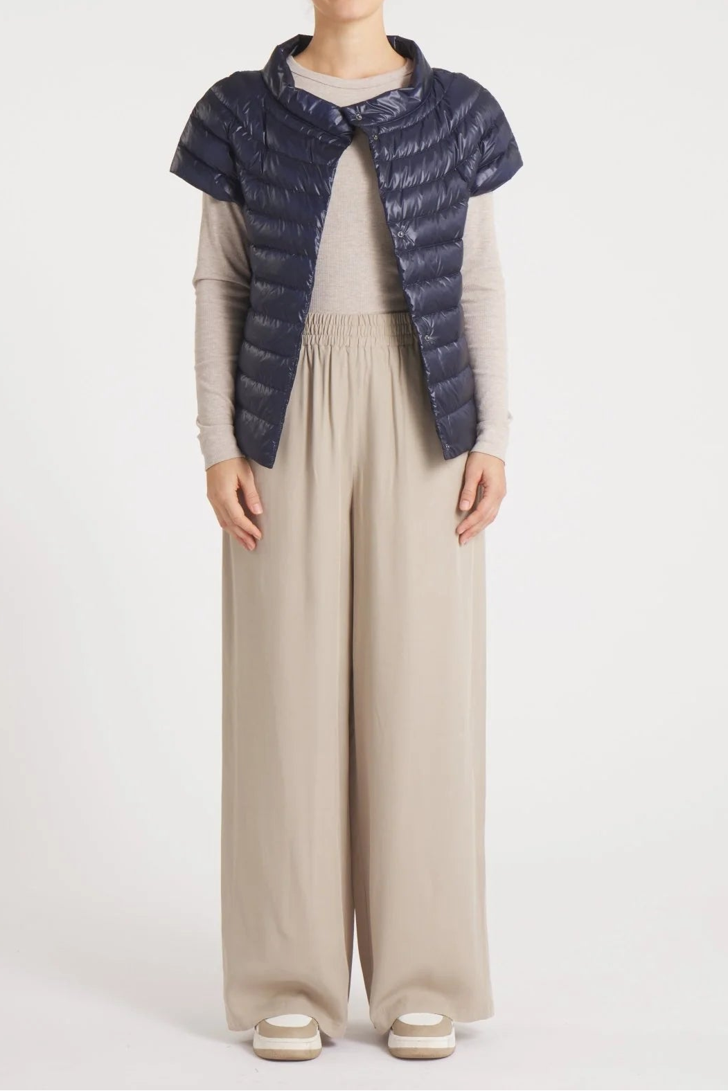 A model stands against a light gray studio background wearing a dark navy quilted puffer vest with a high collar over a beige long-sleeve top and wide-leg beige trousers.