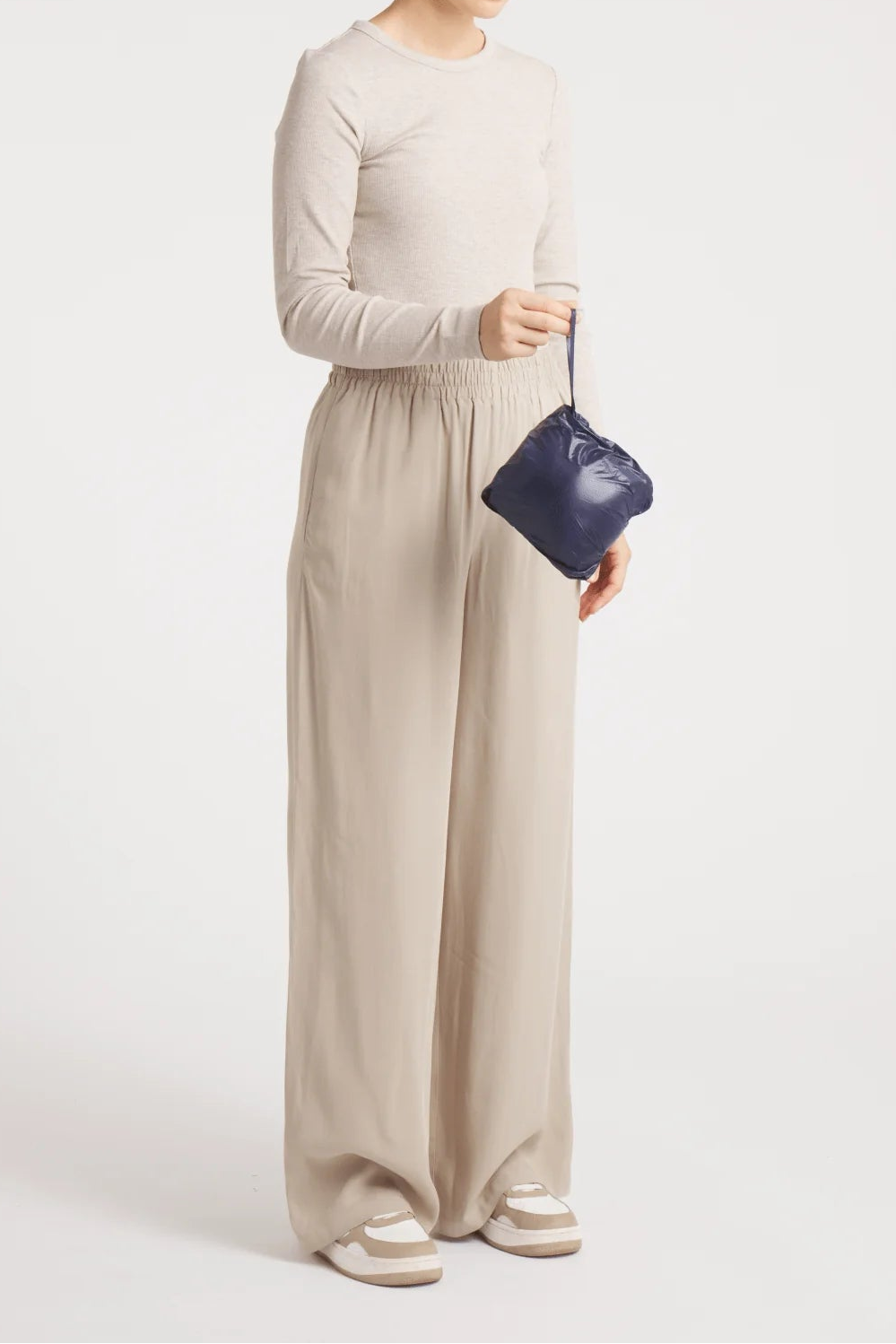 A woman modeling a light beige long-sleeve top and matching wide-leg trousers, holding a small dark blue handbag against a plain light gray studio background.