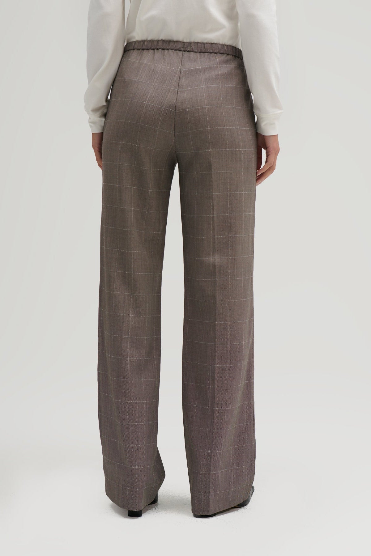 A person shown from the back wearing high-waisted, wide-leg trousers in a muted taupe-brown color with a subtle windowpane plaid pattern, paired with a white long-sleeve top.