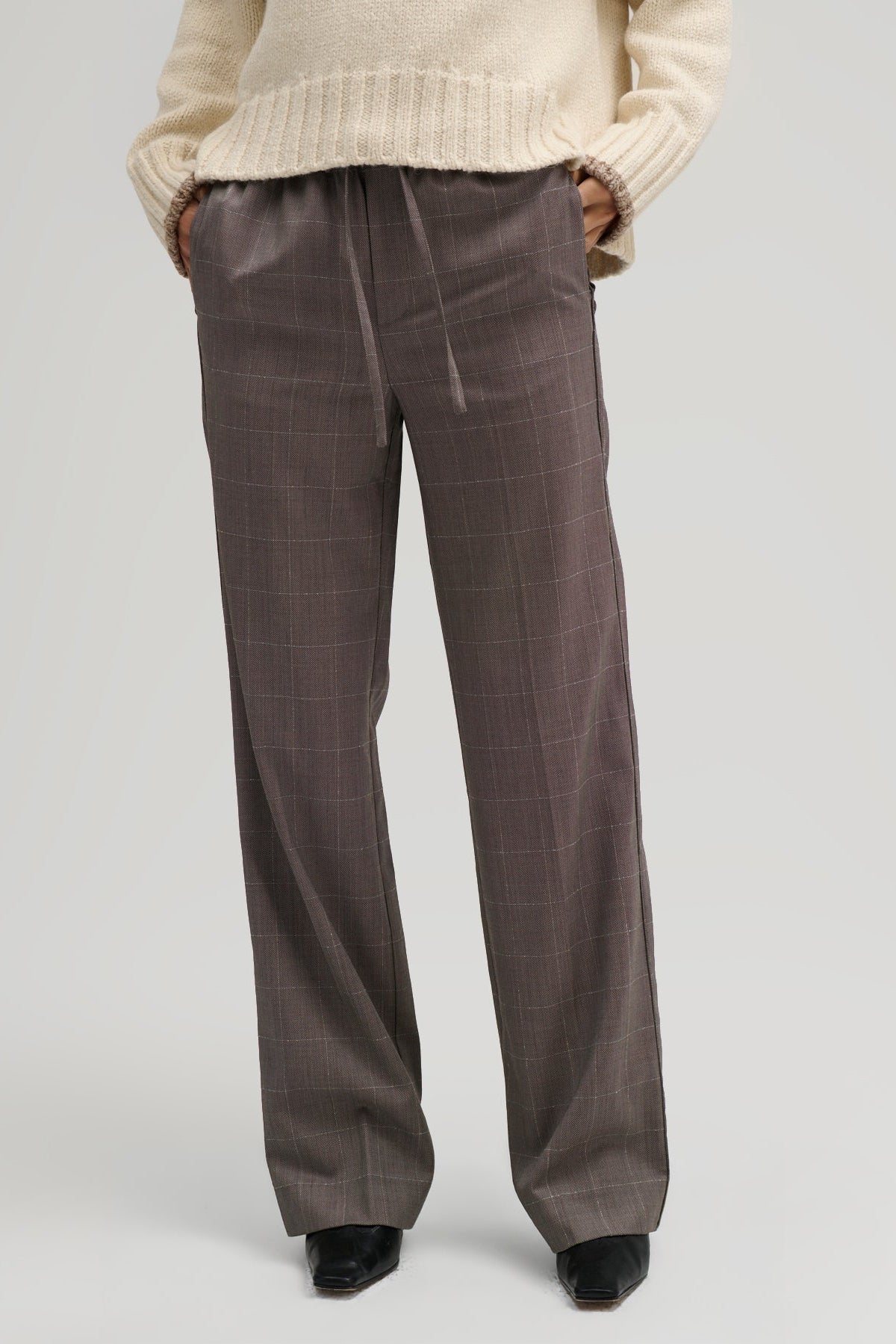 A person wearing wide-leg brown plaid trousers with a drawstring waist, paired with a cream knit sweater and black shoes, shown from the waist down on a light gray studio background.