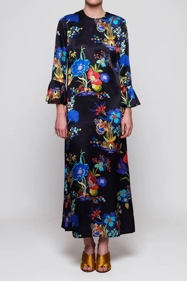 A model stands against a light gray studio background wearing a long-sleeved midi dress with a vibrant multicolor floral print on a black base, paired with metallic gold sandals.