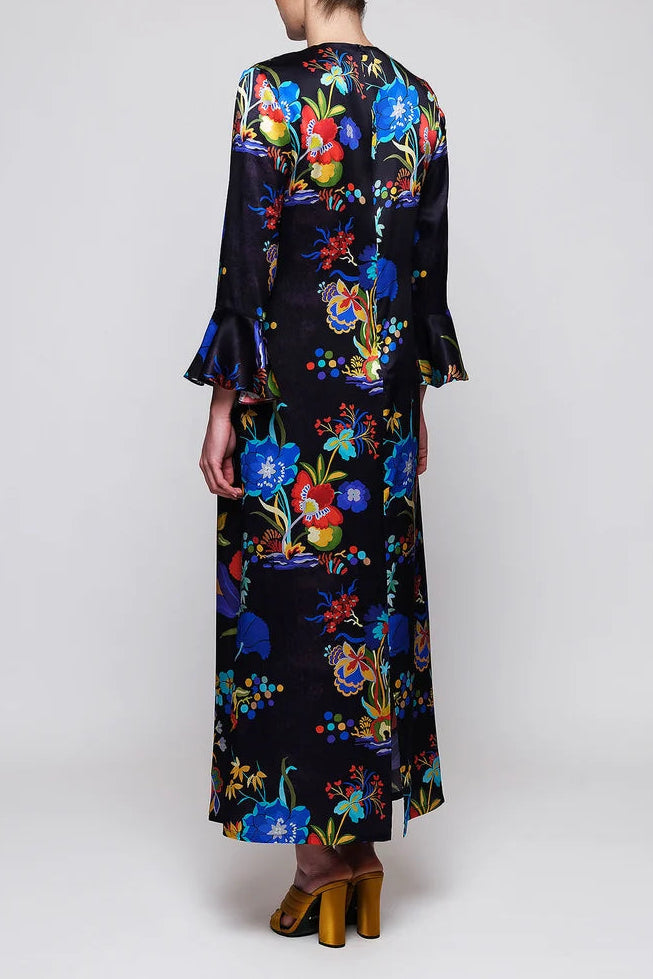 A woman viewed from the back wearing a long-sleeved maxi dress with a vibrant multicolor floral print on a black base, paired with brown block-heel sandals, against a light gray studio background.