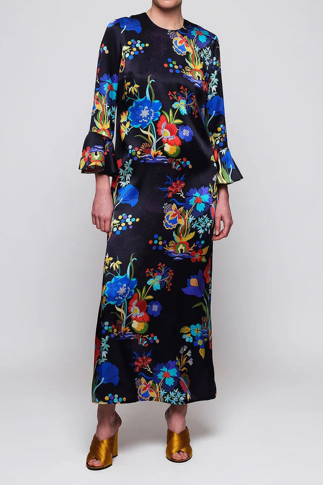 A model wearing a long-sleeved midi dress with a vibrant multicolor floral print on a black satin-like fabric, styled with gold metallic sandals and black sunglasses against a light gray studio background.