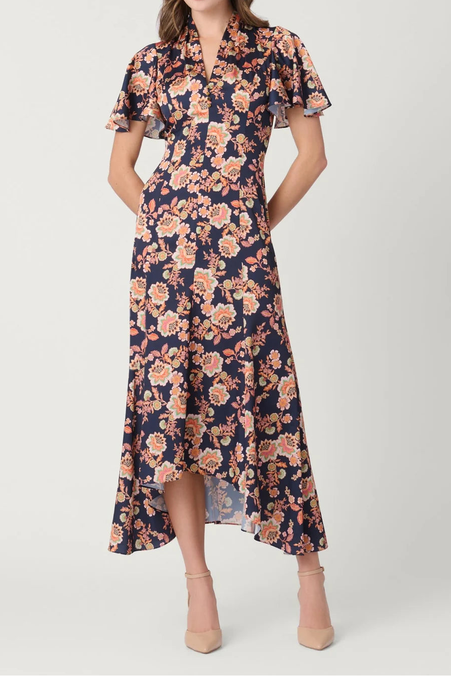 A woman models a navy blue midi dress with a pink and orange floral print, featuring short flutter sleeves, a high collar, and a tiered ruffled hemline, paired with beige high heels against a light gray studio background.