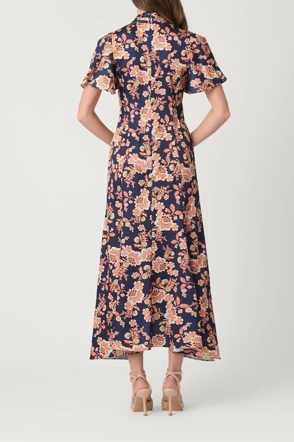 A woman viewed from the back wearing a navy midi dress with a pink and peach floral print, paired with beige high-heeled sandals, against a light gray studio background.