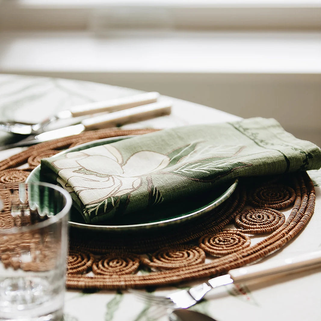 A styled table setting featuring a folded sage green linen napkin with a white floral print, layered over a dark green plate and a brown woven placemat.