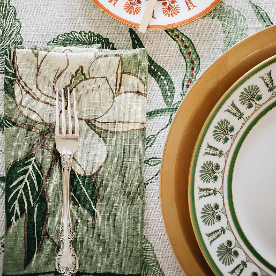 A styled tabletop setting featuring a sage green linen napkin with a large cream magnolia floral print, folded over on a matching patterned tablecloth. A silver-toned ornate fork rests on the napkin, next to stacked plates including a gold charger and white plates with green botanical borders.