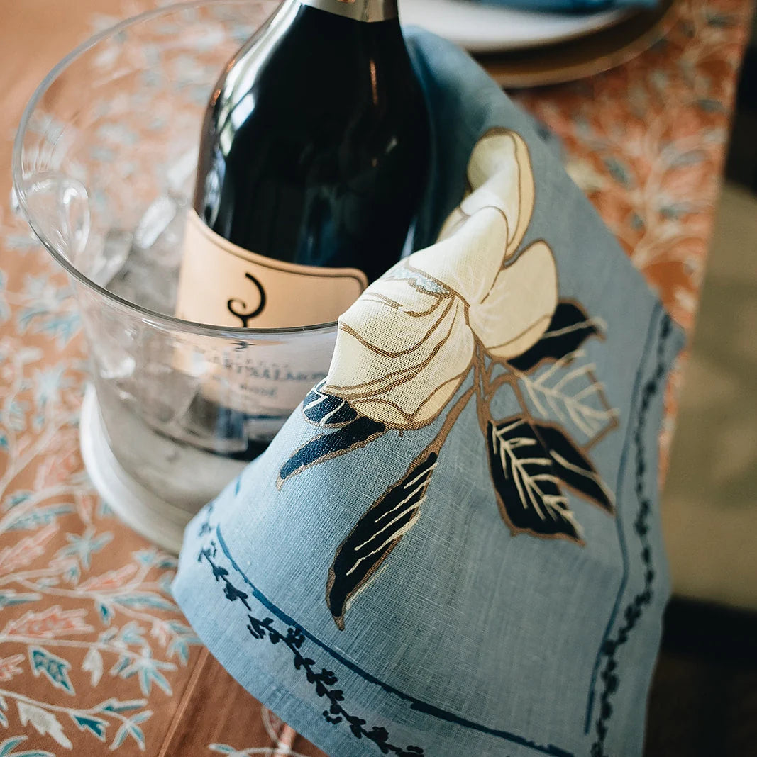 A light blue linen napkin with embroidered white, black, and gold magnolia flowers, draped over a dark wine bottle in a clear glass ice bucket on a patterned tablecloth.