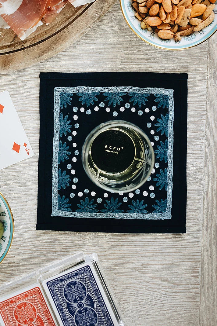 A square black napkin with a blue and white floral pattern and a clear glass resting on it, styled in a flat-lay arrangement with playing cards, olives, nuts, and cured meat on a light wood surface.