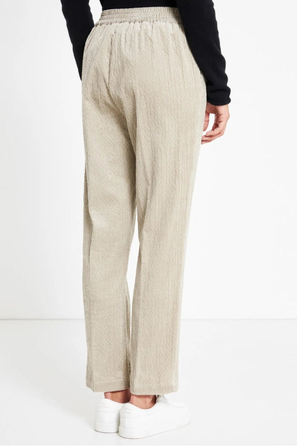 A person shown from the back wearing light beige textured wide-leg trousers, a black long-sleeve top, and white sneakers against a clean white studio background.