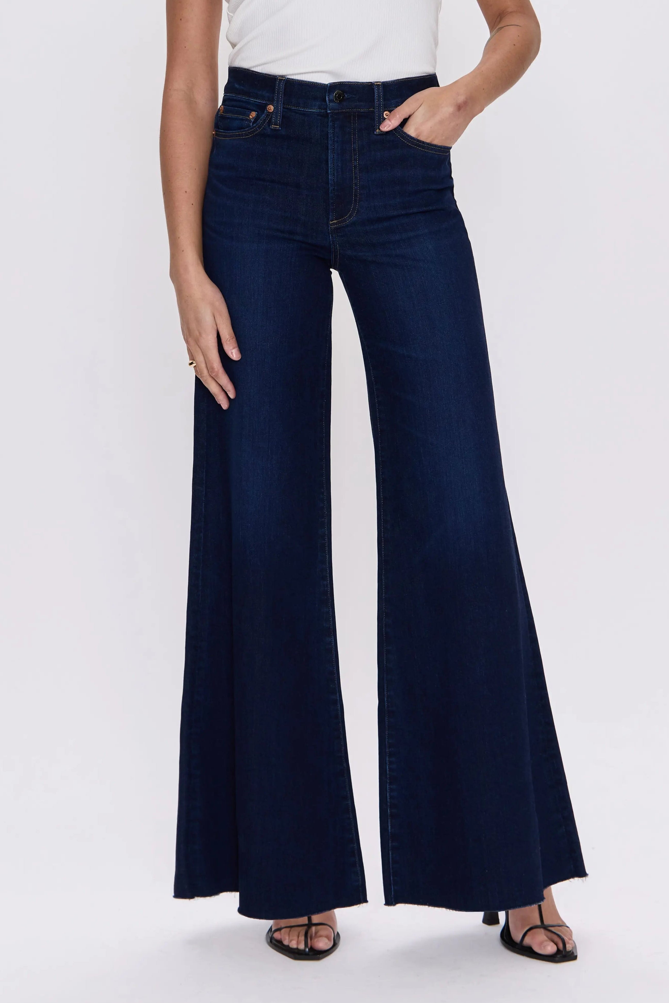 A model wearing dark blue wide-leg flare jeans with a white tank top, standing against a light gray studio background.