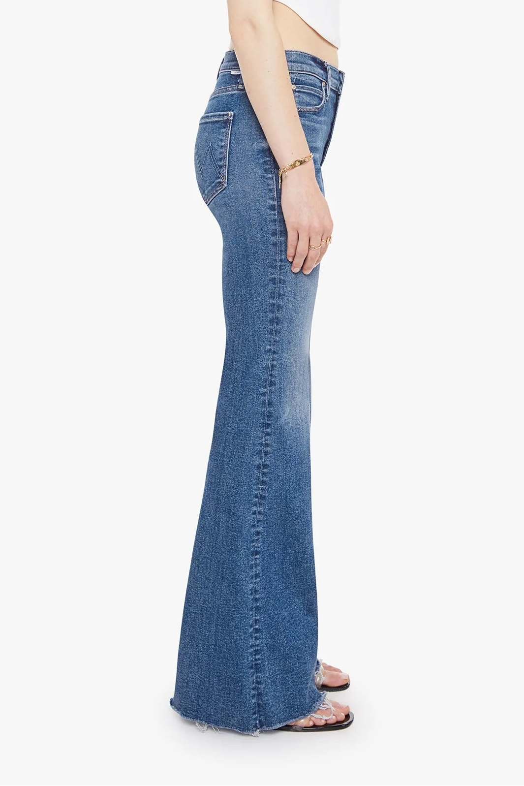 A side-profile view of a model wearing blue high-waisted flare jeans, a white crop top, and sandals against a plain white background.