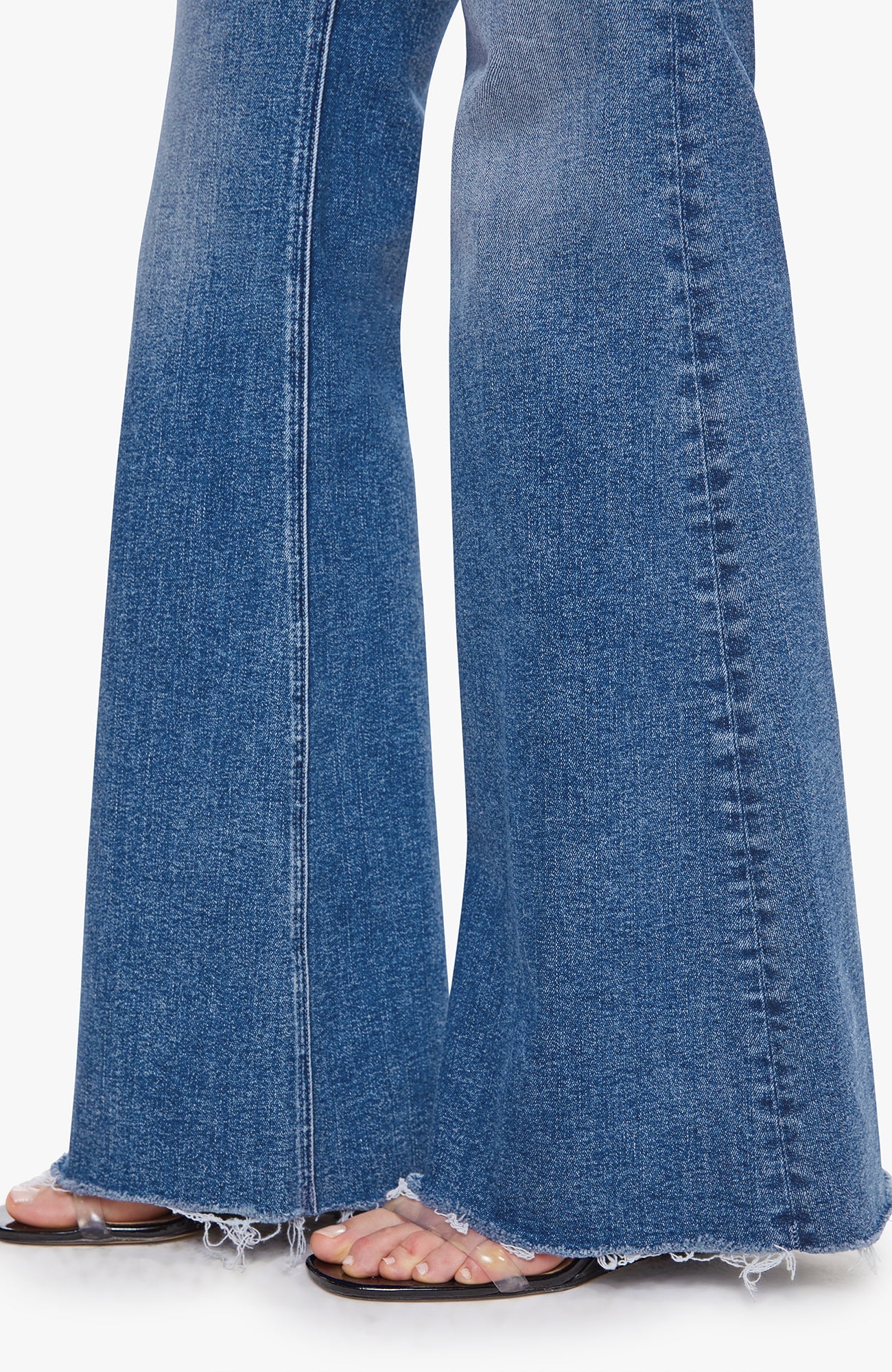 Close-up of a person wearing blue flare jeans with frayed hems and clear flat sandals, shown against a plain light background.