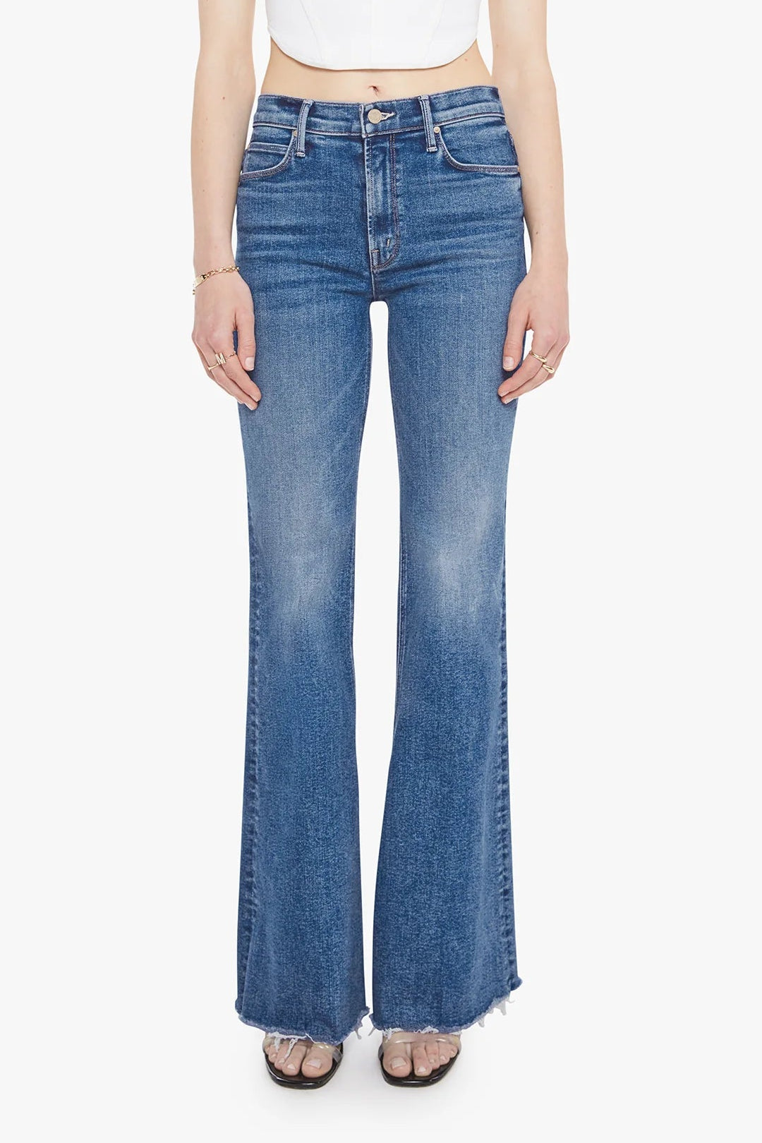 A model wearing a pair of medium-wash blue flare jeans with a frayed hem, paired with a white crop top and flat sandals, against a plain white background.