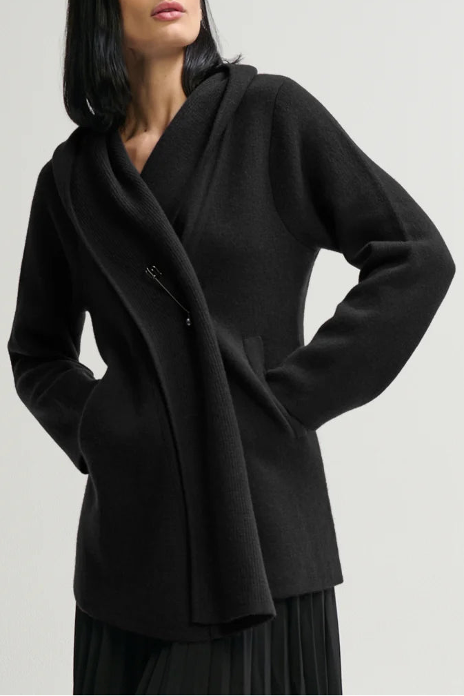 A woman modeling a black wrap-style coat with a draped collar and front zipper, paired with a black pleated skirt, against a plain light gray studio background.