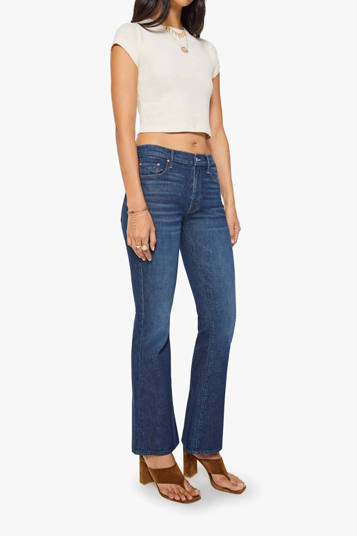 A woman modeling a cream short-sleeve cropped top and blue high-waisted straight-leg jeans with brown block-heel sandals, set against a plain light gray studio background.