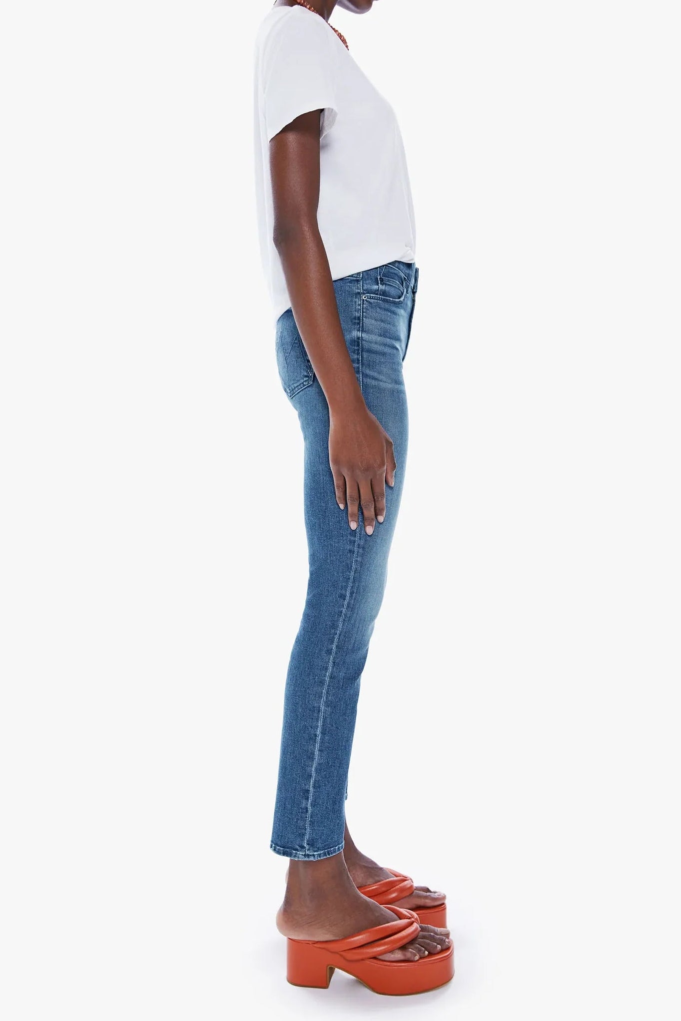 A model shown in profile wearing blue skinny jeans, a white short-sleeve t-shirt, and orange platform mule sandals against a plain light gray background.