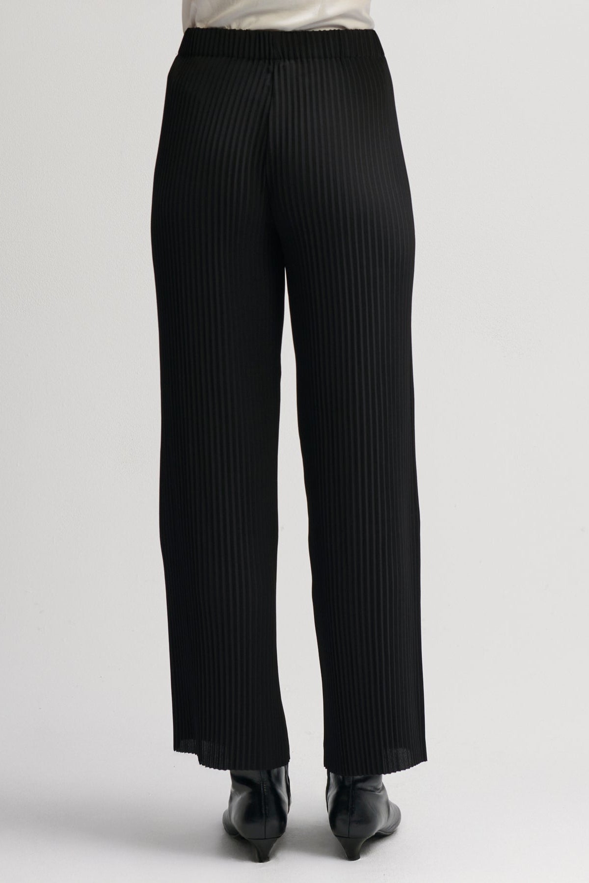 Black pleated wide-leg trousers shown from the back on a model, paired with black pointed-toe heels against a light gray studio background.