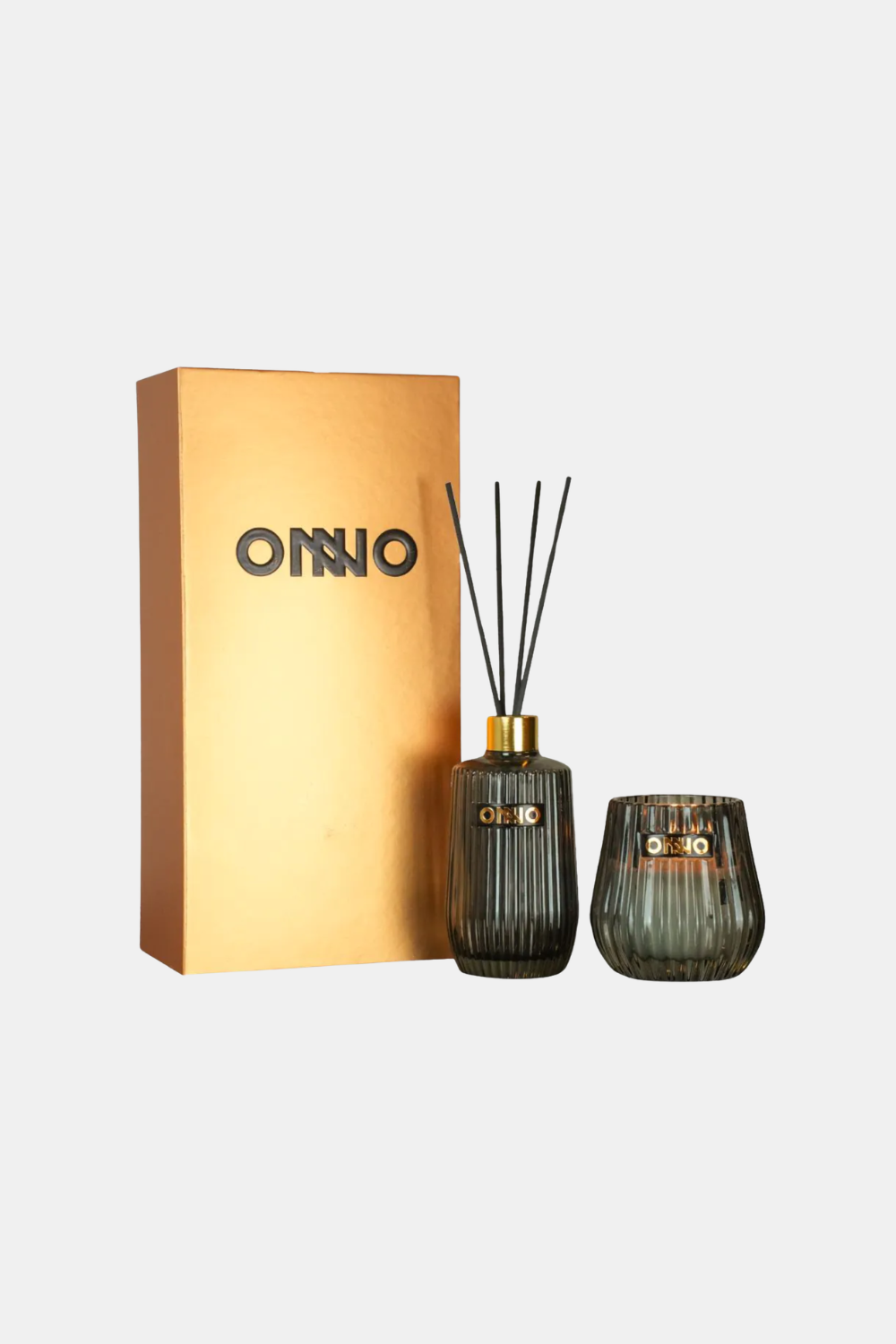 An ONNO reed diffuser set with a gold-colored box, a dark ribbed glass reed diffuser bottle with reeds, and a matching dark ribbed glass candle holder, shown on a light gray background.