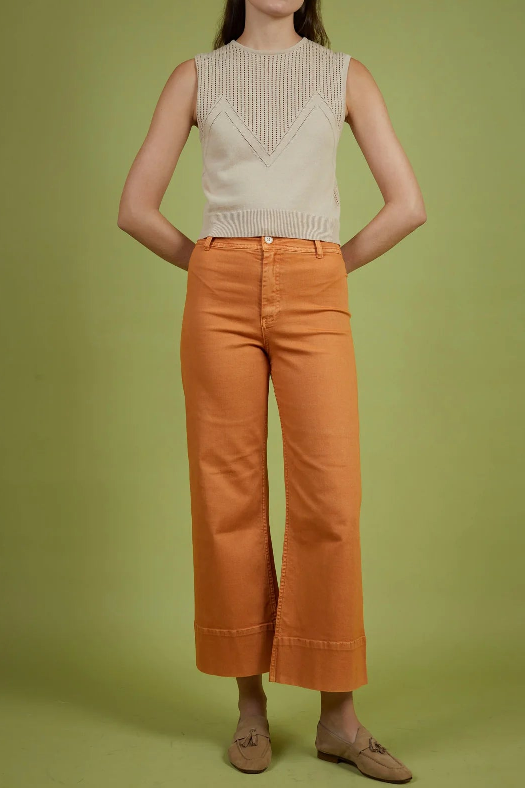 Woman wearing a beige sleeveless top and orange pants against a green background