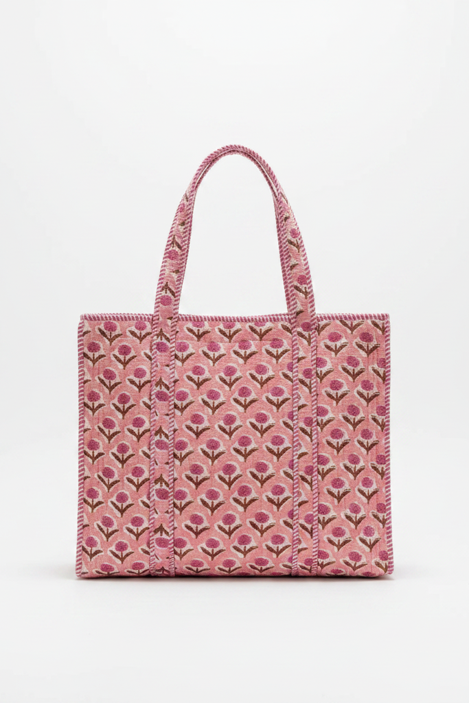 A pink tote bag with a repeating floral block-print pattern sits on a rustic wooden table in a bright, modern living room setting.