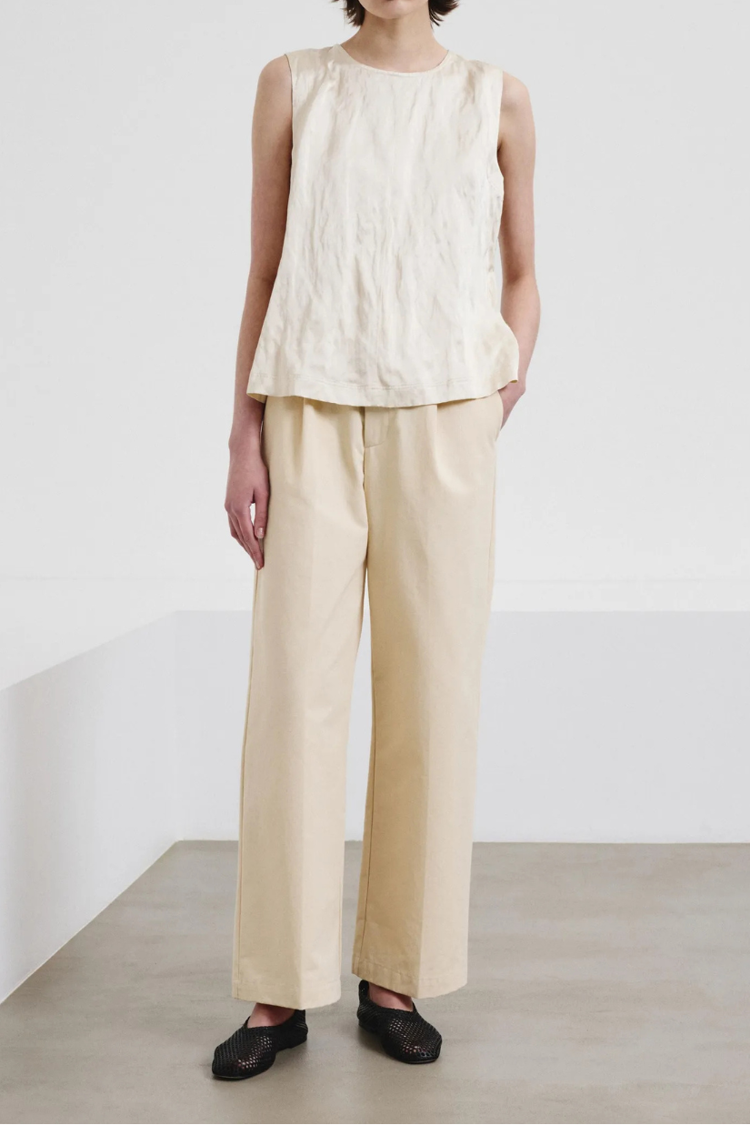 A model wearing a cream-colored sleeveless top and matching wide-leg trousers, styled with black flat shoes and sunglasses in a minimalist studio setting.