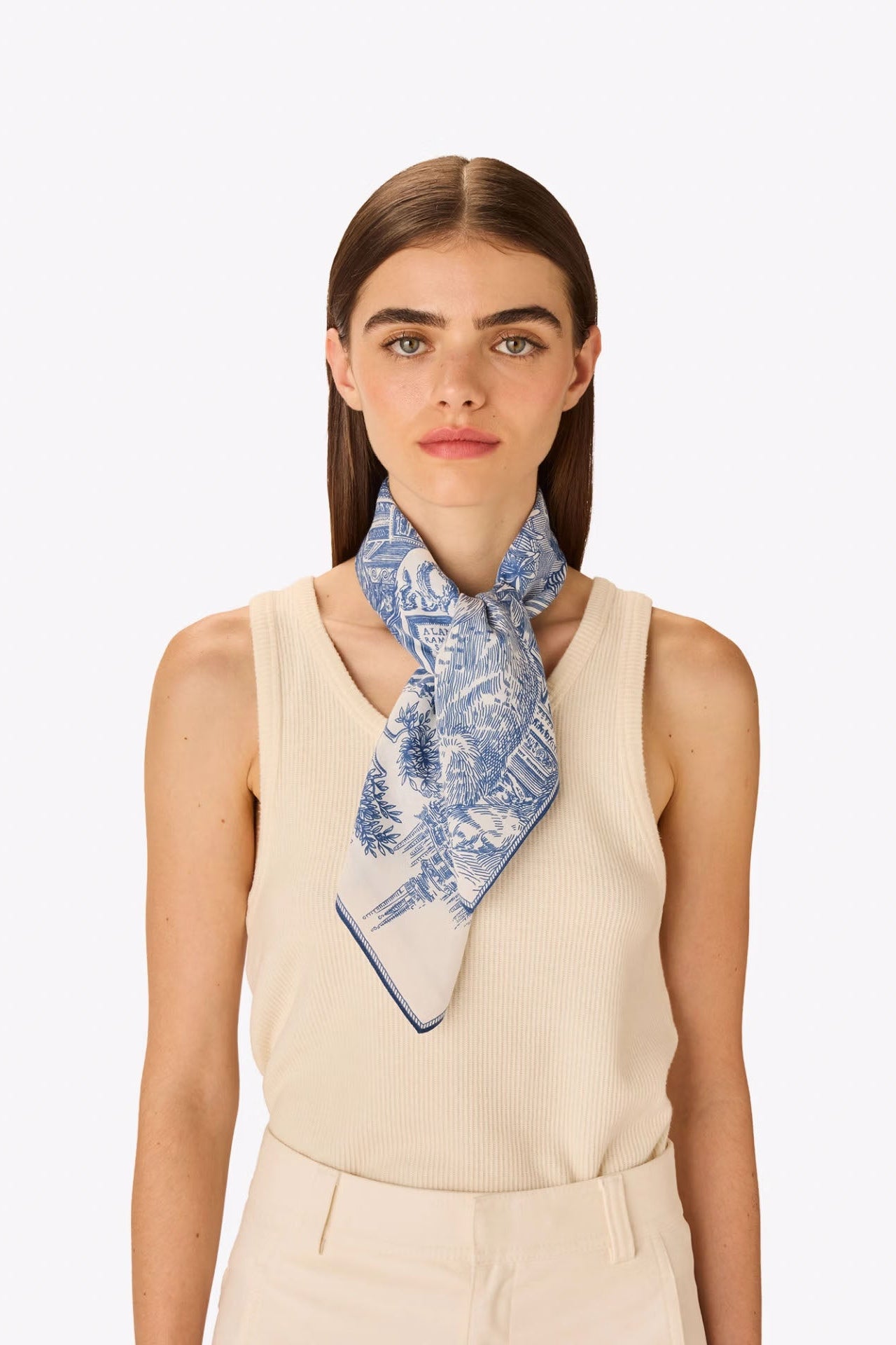 A woman modeling a blue and white patterned silk scarf tied around her neck, paired with a cream ribbed sleeveless top against a light gray studio background.