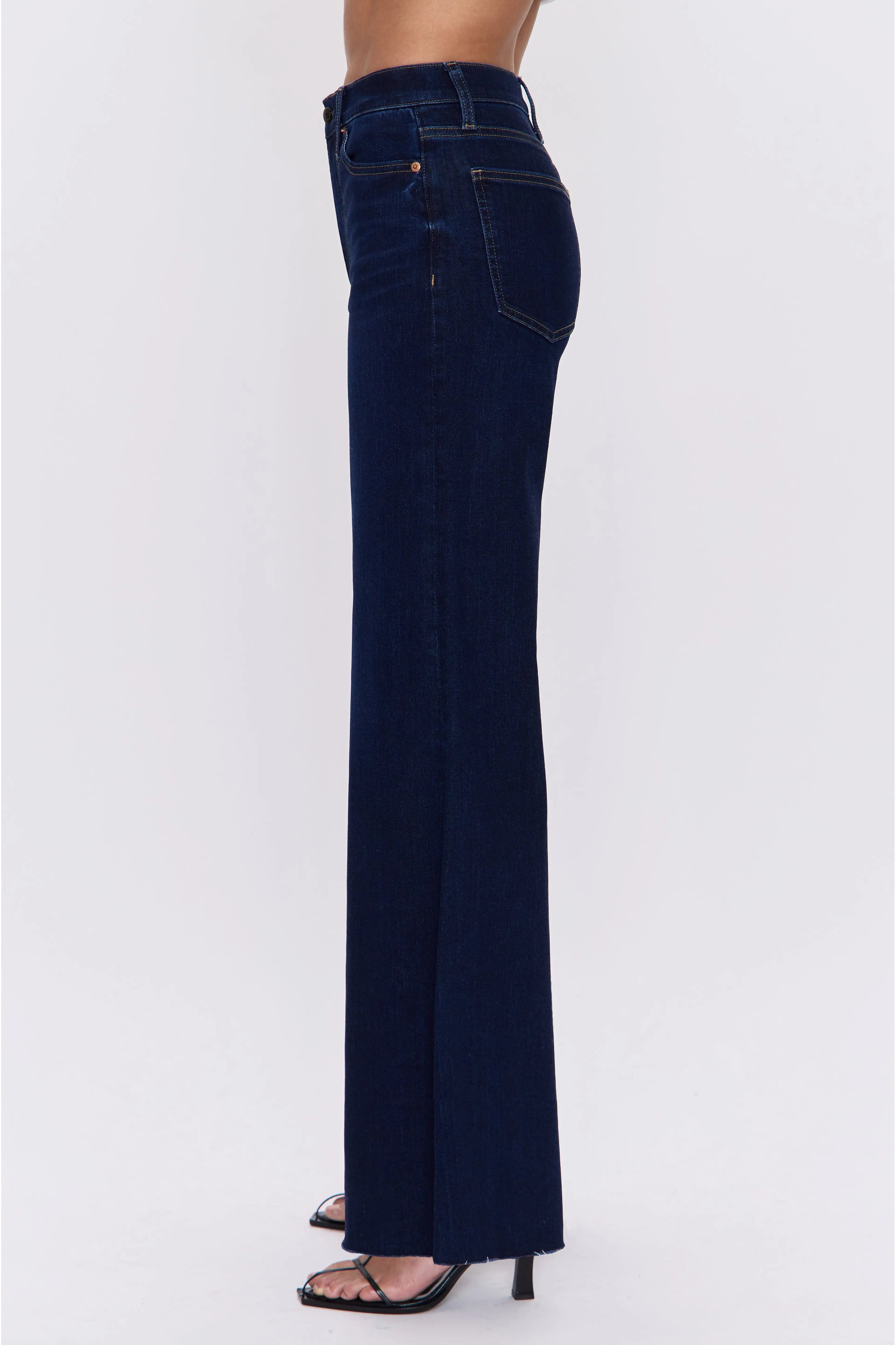 A side profile view of a model wearing dark navy blue flare jeans and black high-heeled sandals against a plain white studio background.