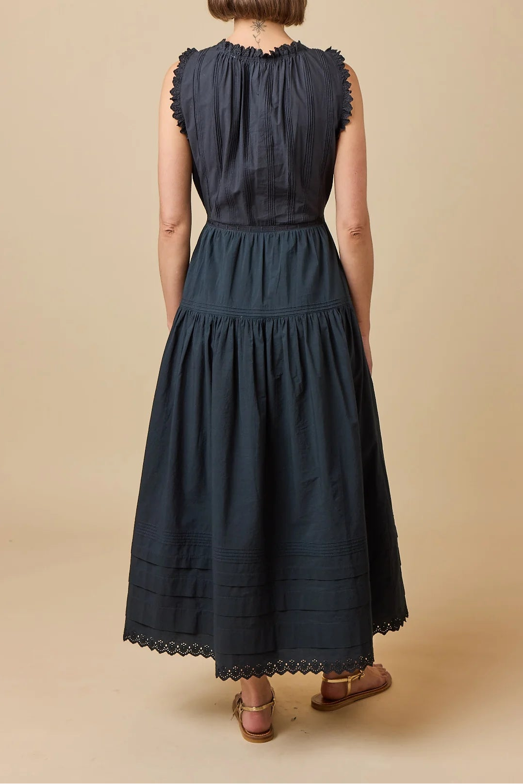 A woman viewed from the back wearing a sleeveless dark navy midi dress with a gathered waist and scalloped lace trim, standing against a plain beige studio background.