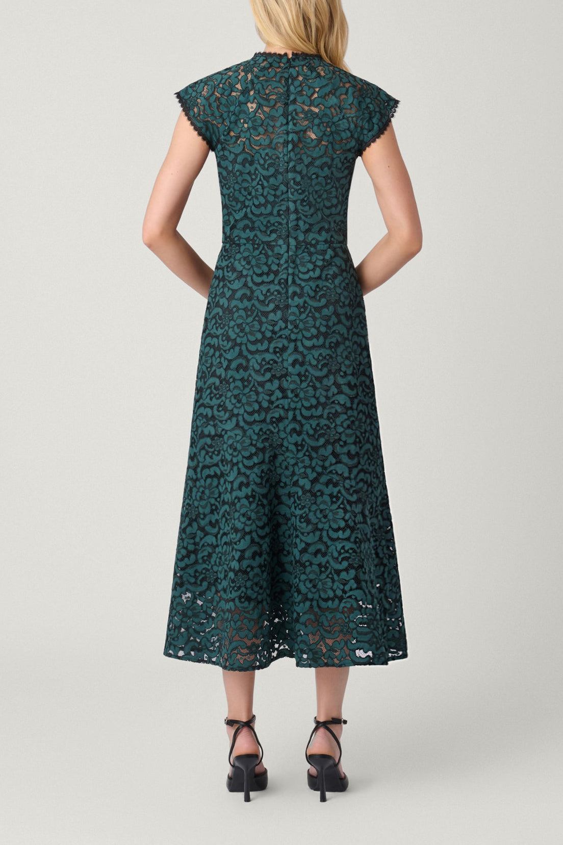 A woman viewed from the back wearing a dark teal lace midi dress with cap sleeves and black heels, standing against a light gray studio background.