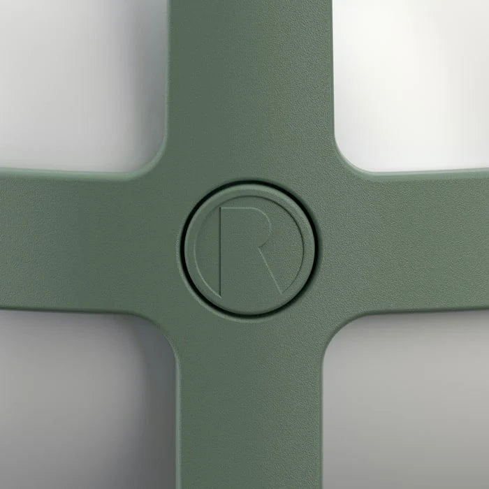 Close-up of a sage green cross-shaped product with rounded corners and a circular embossed letter 'R' in the center of the vertical bar, set against a light gray background.