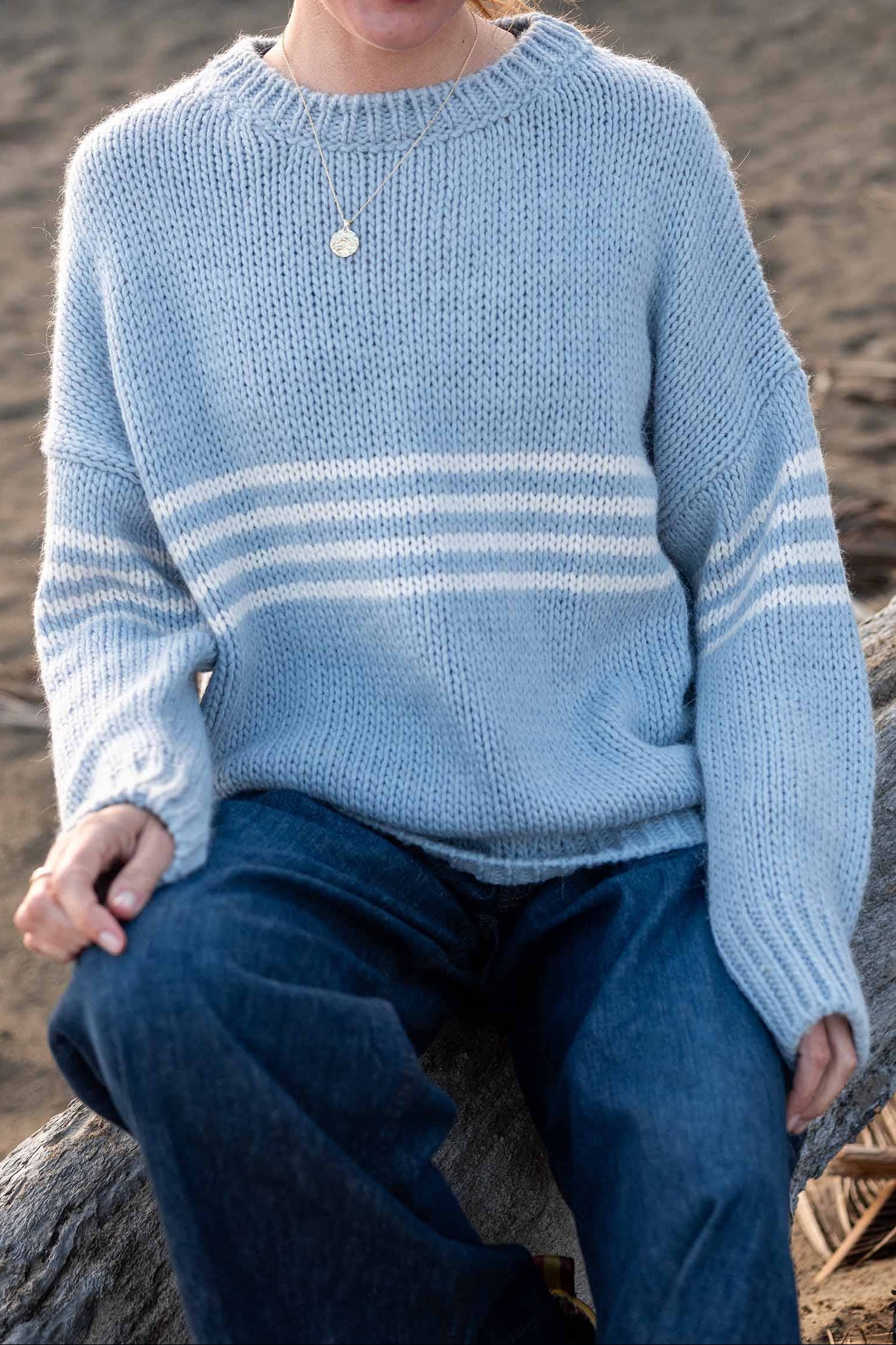 Person wearing a light blue sweater with white stripes sitting on a log by a sandy area.