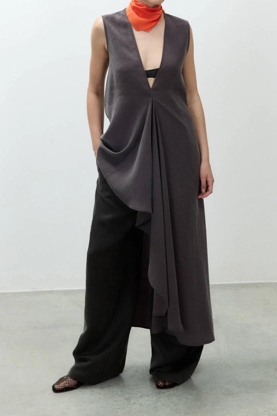 A model stands in a minimalist studio wearing a dark gray draped sleeveless top with a deep V-neck, wide-leg black pants, and a bright orange neck scarf.