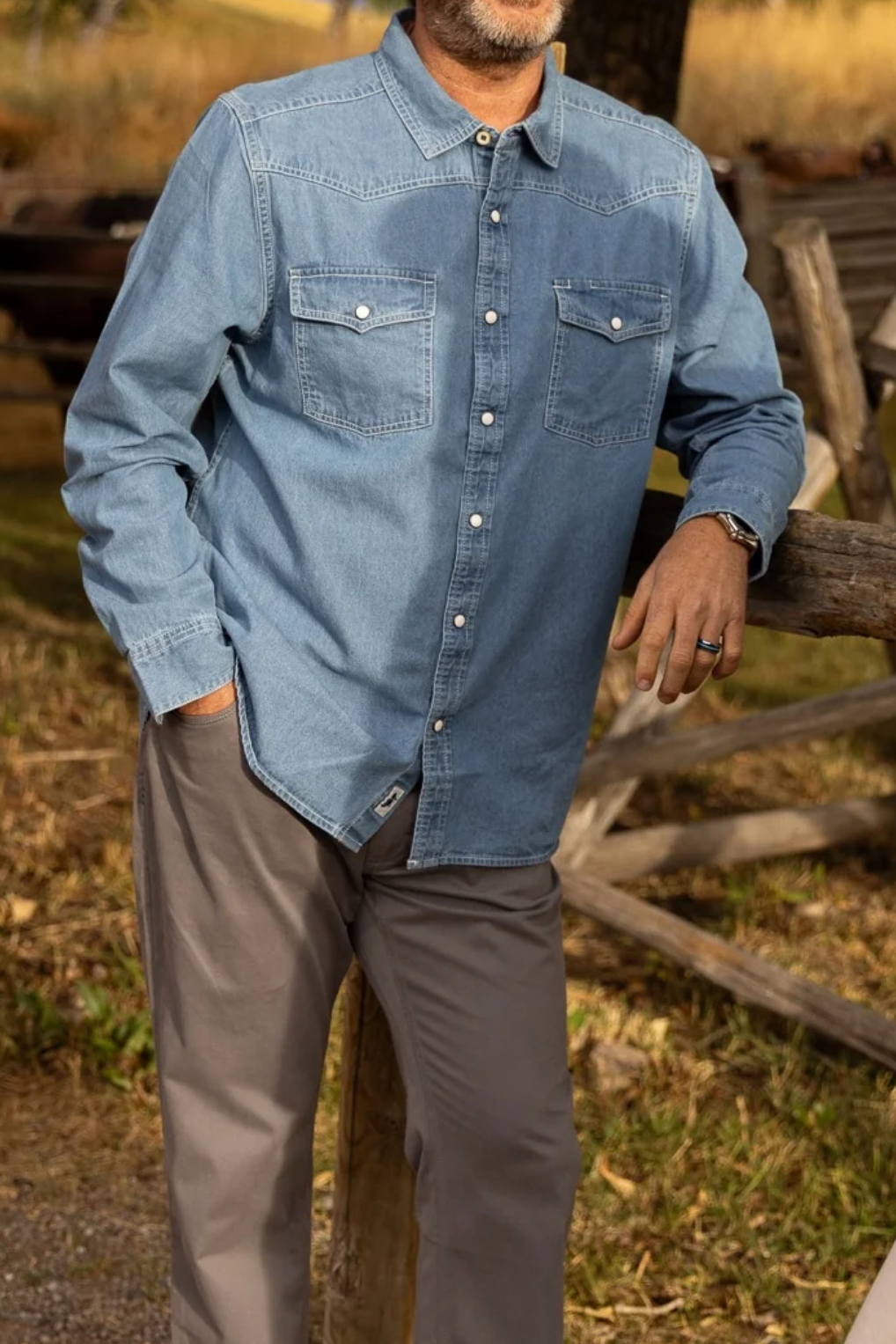 Man wearing a blue denim shirt standing in a rustic outdoor setting with wooden fences and trees.