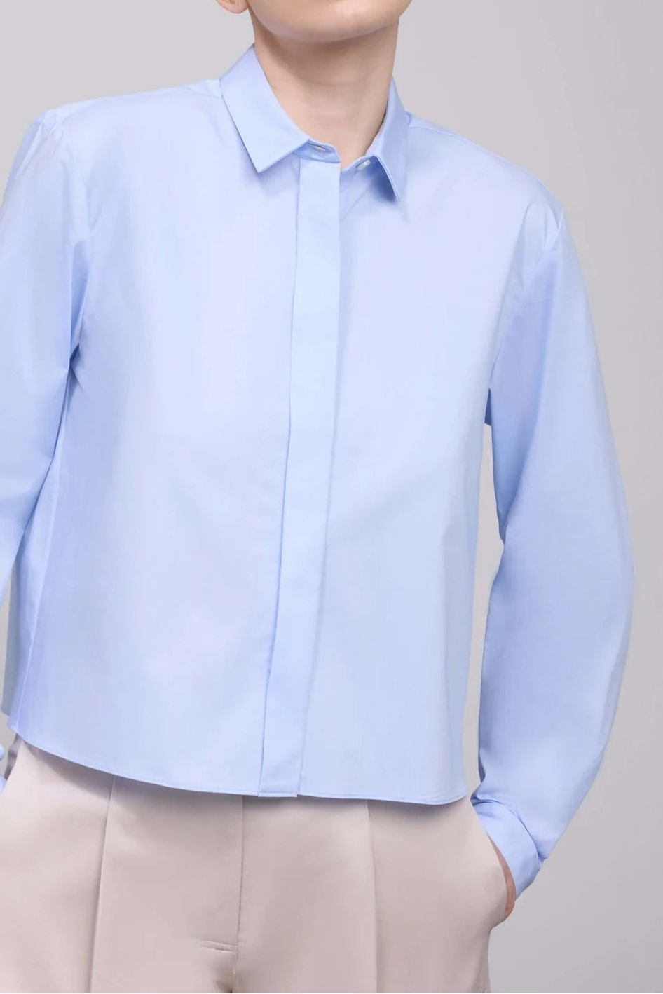 A woman wearing a light blue long-sleeve button-up shirt and beige trousers, standing against a plain light gray studio background.