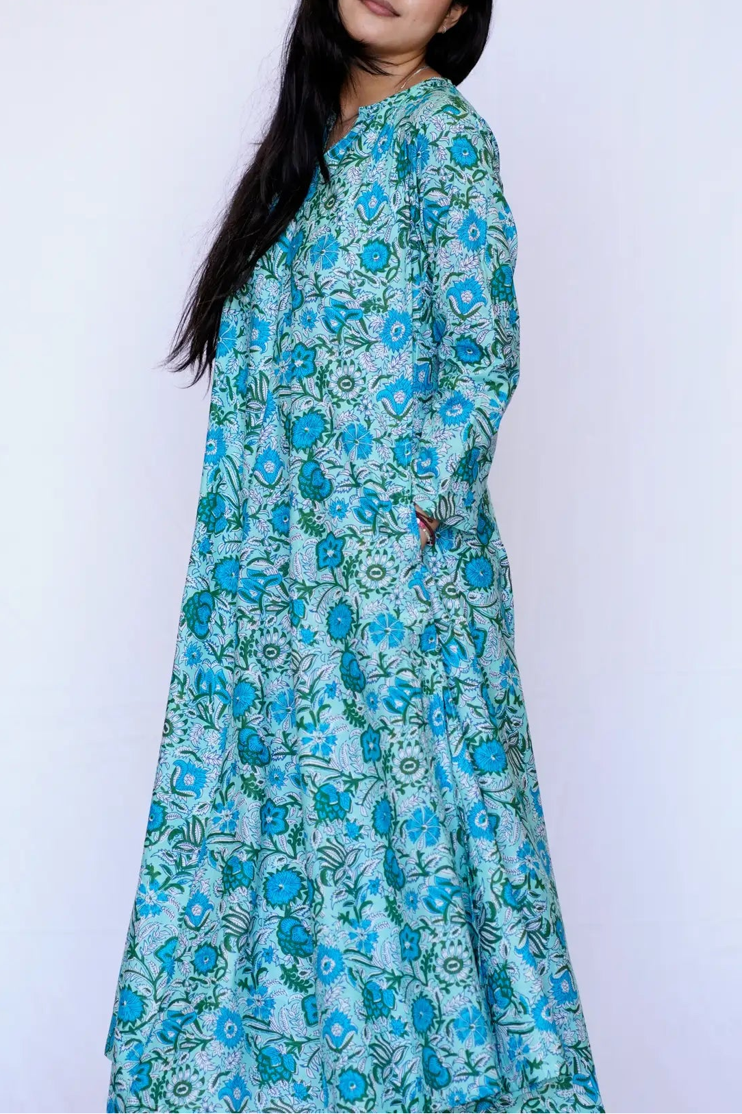 A woman with long black hair is shown from the back wearing a long-sleeved, floor-length dress with a blue and green floral block-print pattern against a plain white background.