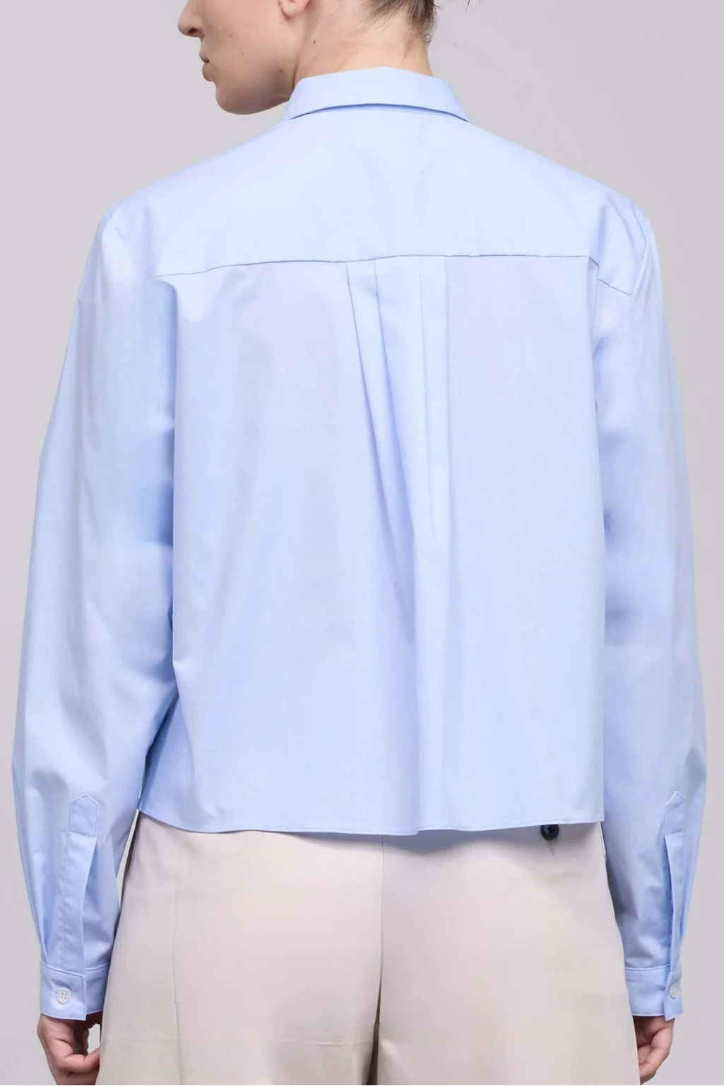 A woman shown from the back wearing a light blue cropped button-up shirt and beige trousers, photographed against a plain light gray studio background.