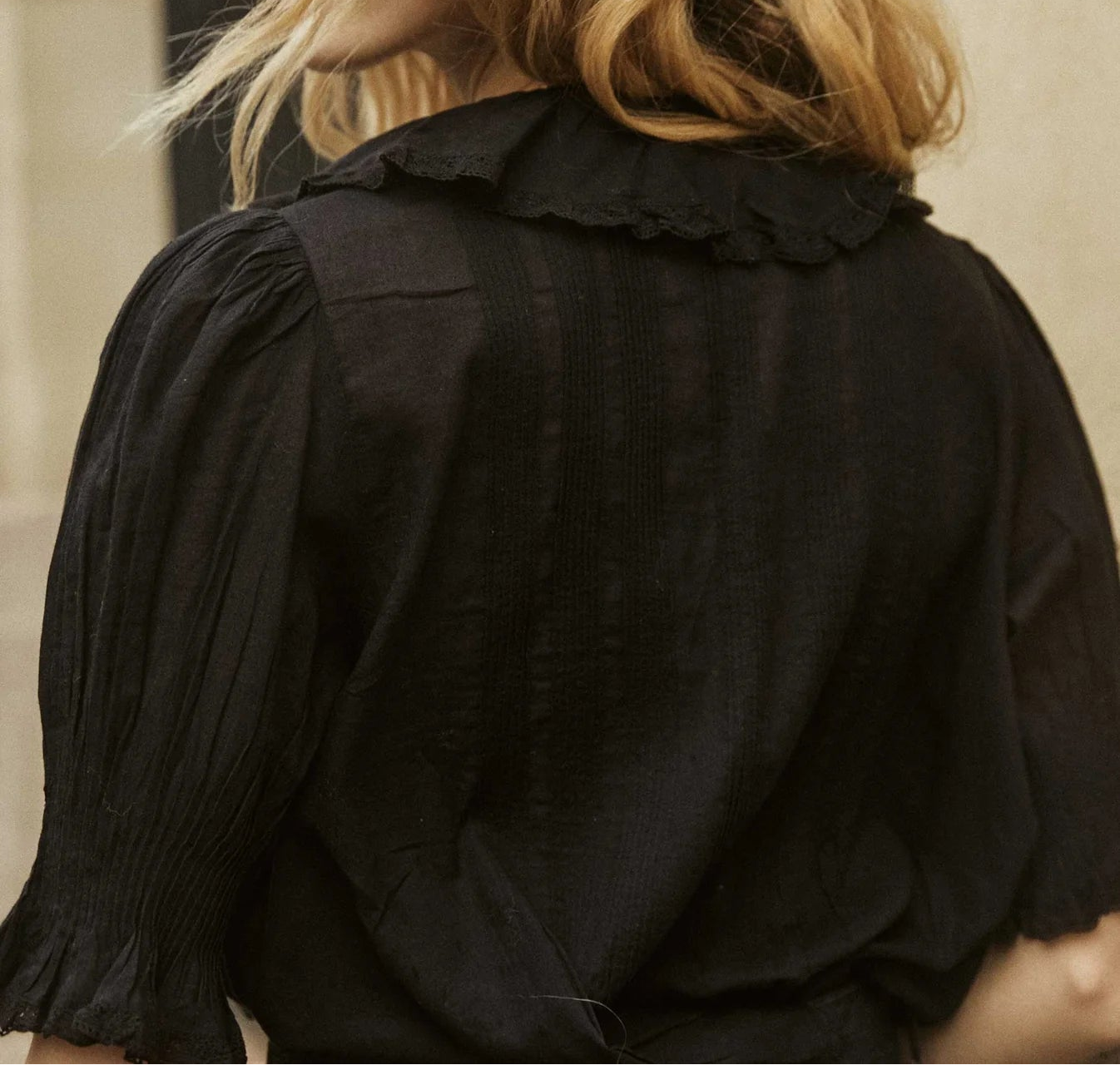 Person wearing a black shirt with a ruffled collar, standing against a neutral background.
