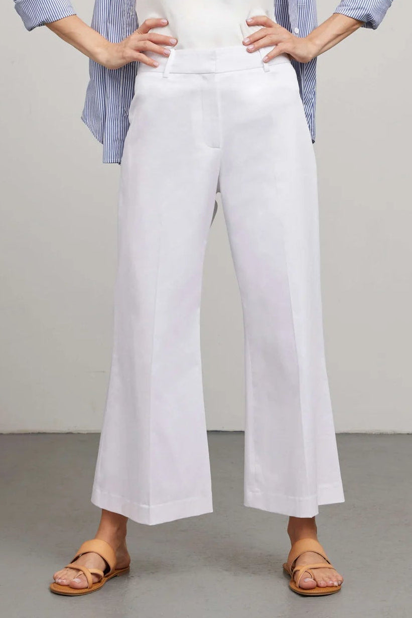 Woman wearing a blue checkered shirt, white top, and white wide-leg pants against a plain background.
