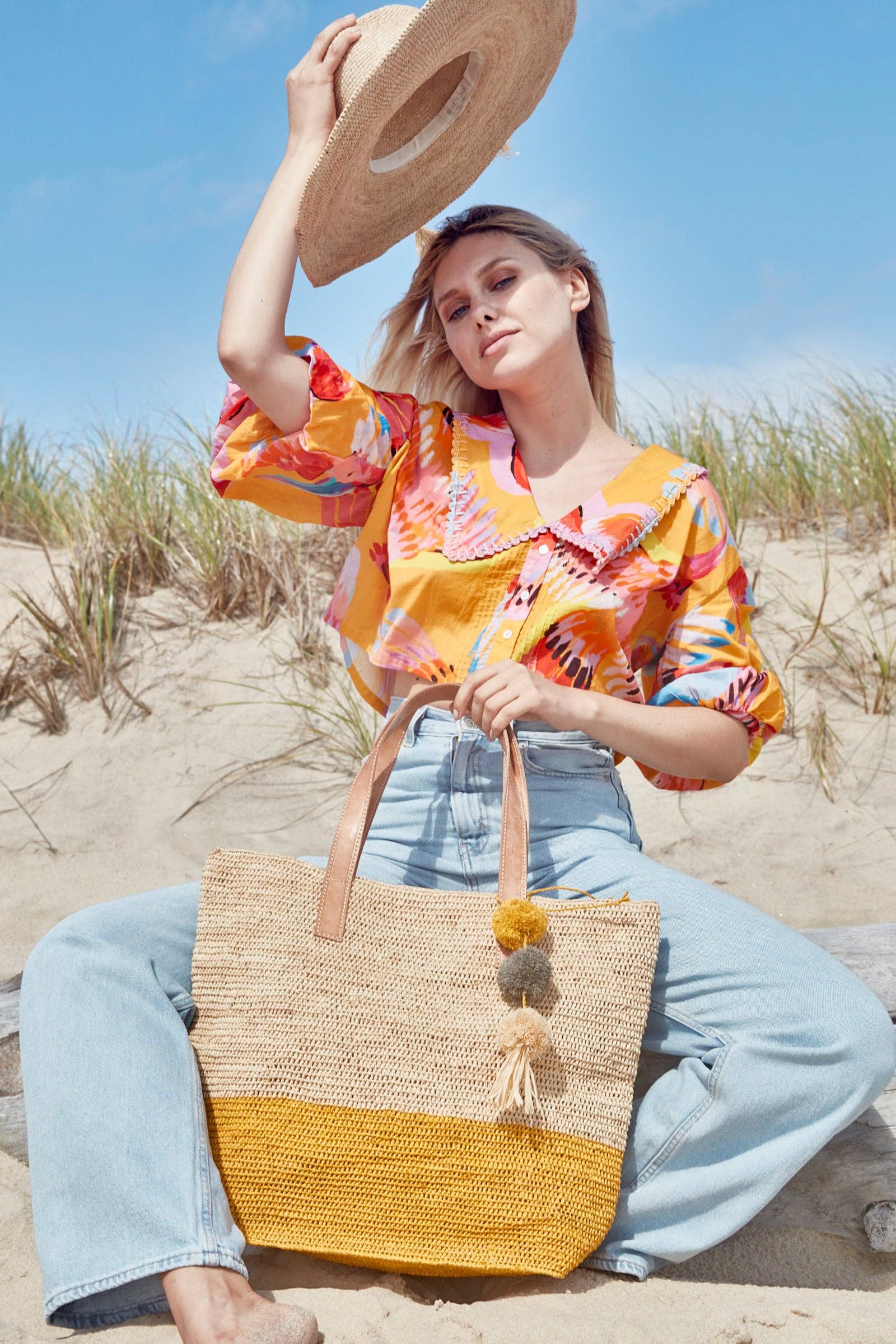 Woman on a beach wearing a colorful shirt and holding a straw bag.