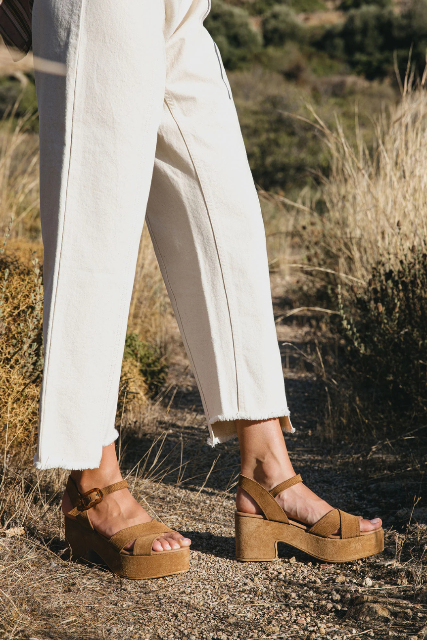 A person wearing off-white cropped pants and tan platform wedge sandals with ankle straps, standing outdoors on a dry grassy path.