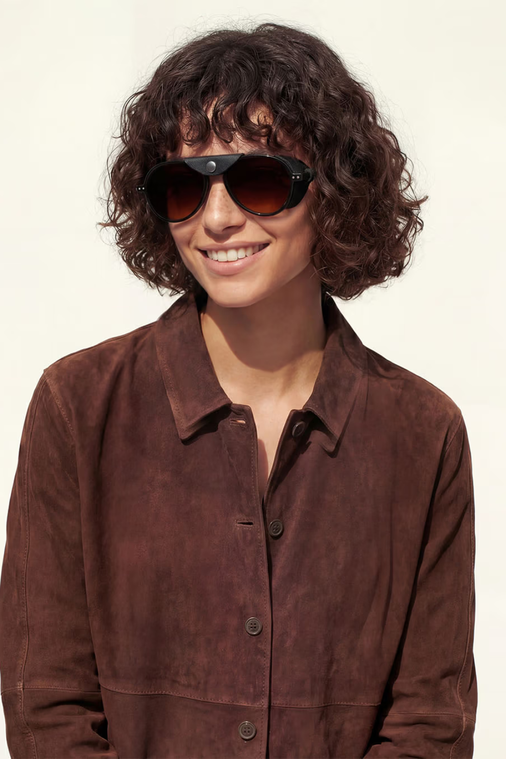 A smiling woman with short curly brown hair wearing dark brown sunglasses and a dark brown suede button-up shirt, photographed against a plain off-white background.