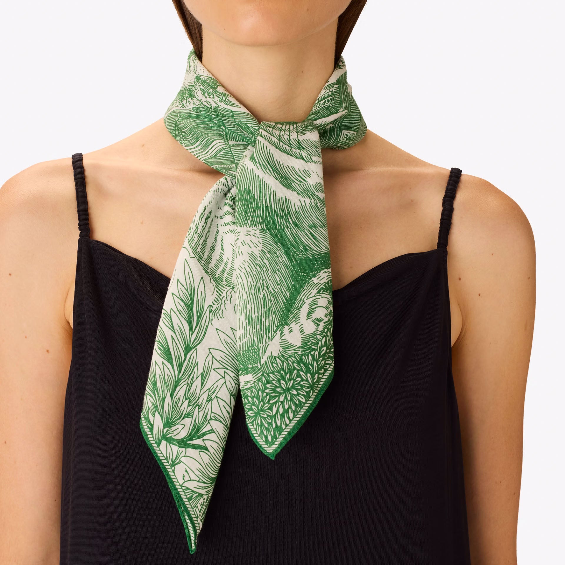 A green and white tropical leaf print silk scarf tied around the neck of a model wearing a black spaghetti strap camisole top, against a plain light gray background.