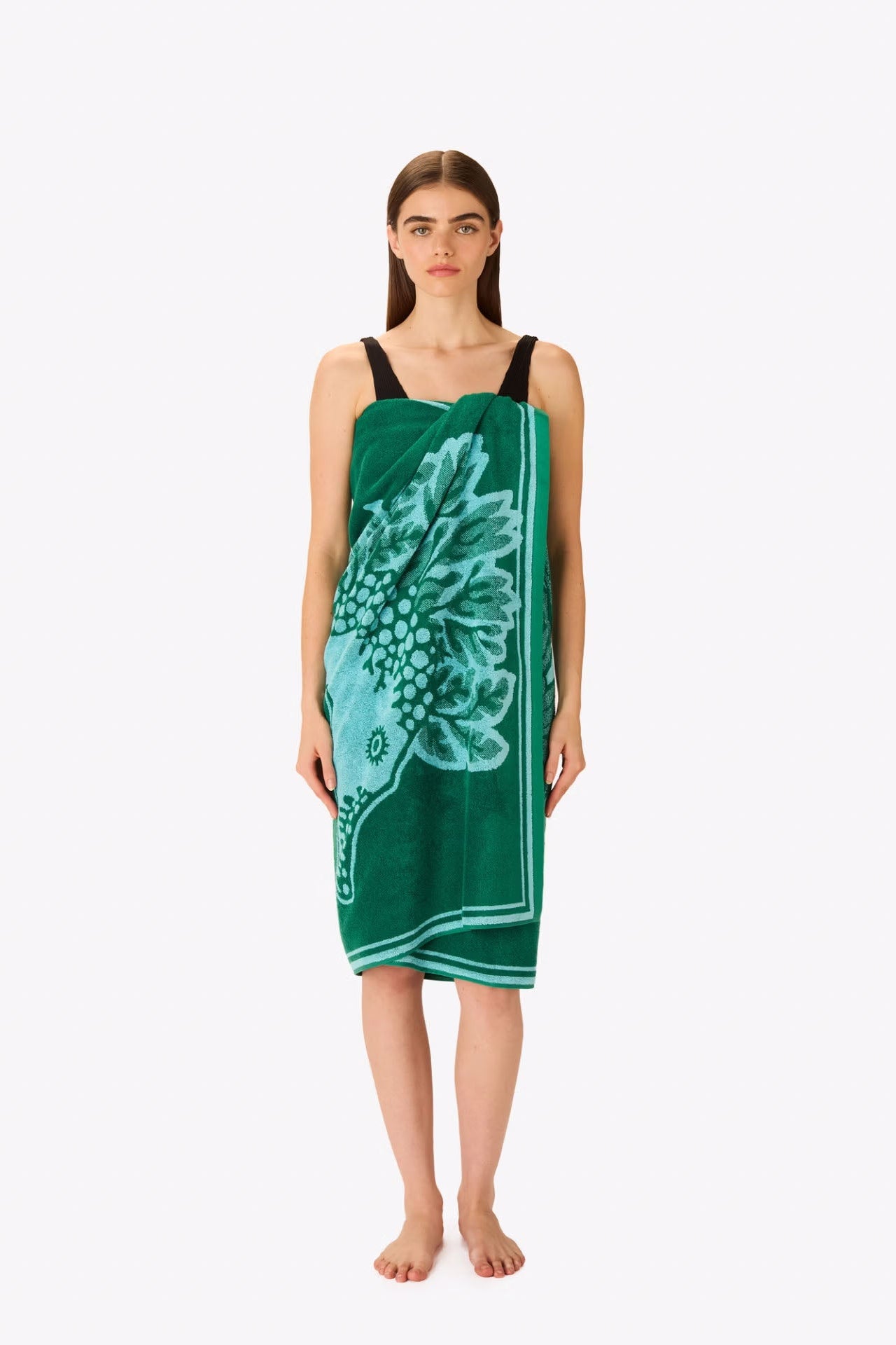 A woman stands barefoot against a light gray studio background wearing a sleeveless green towel dress with a large light blue floral print and thin black shoulder straps.