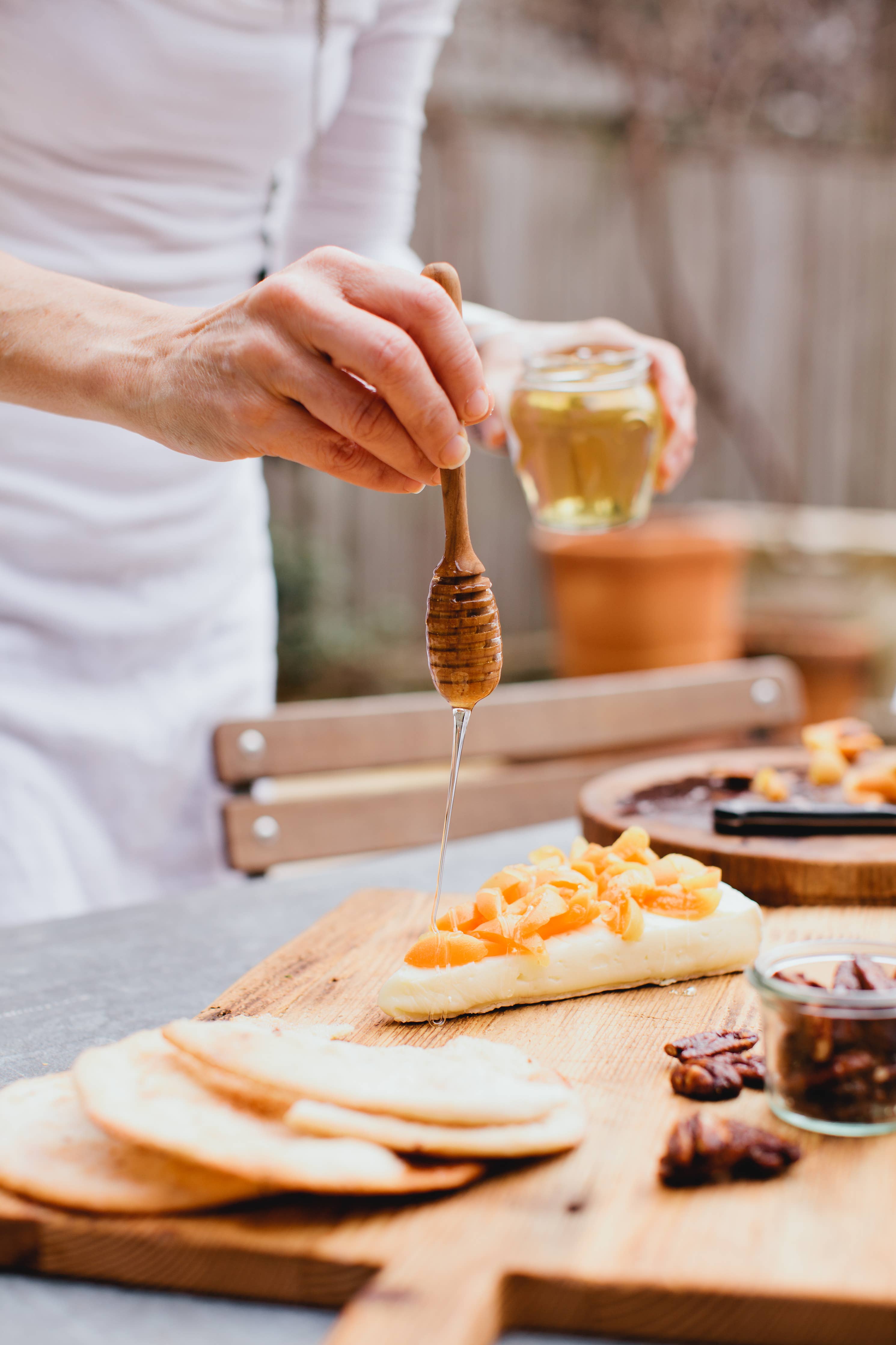 Person drizzling honey from a wooden dipper onto bread with cheese on a wooden board.