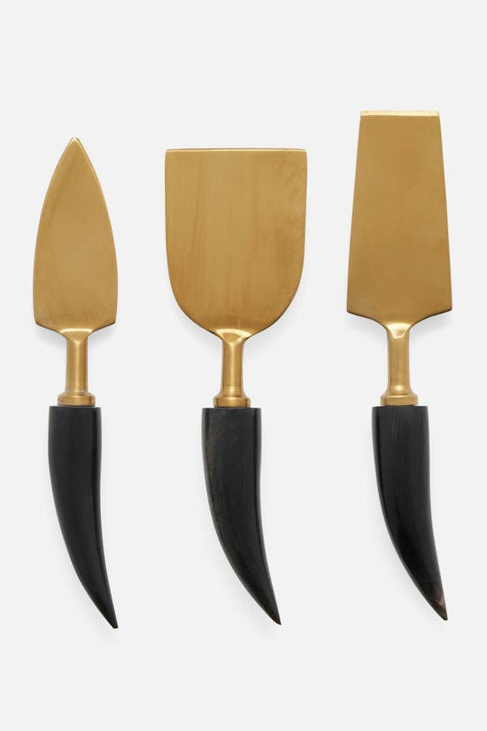 Three cheese knives with gold-toned metal heads and black curved handles, arranged vertically on a light gray background.