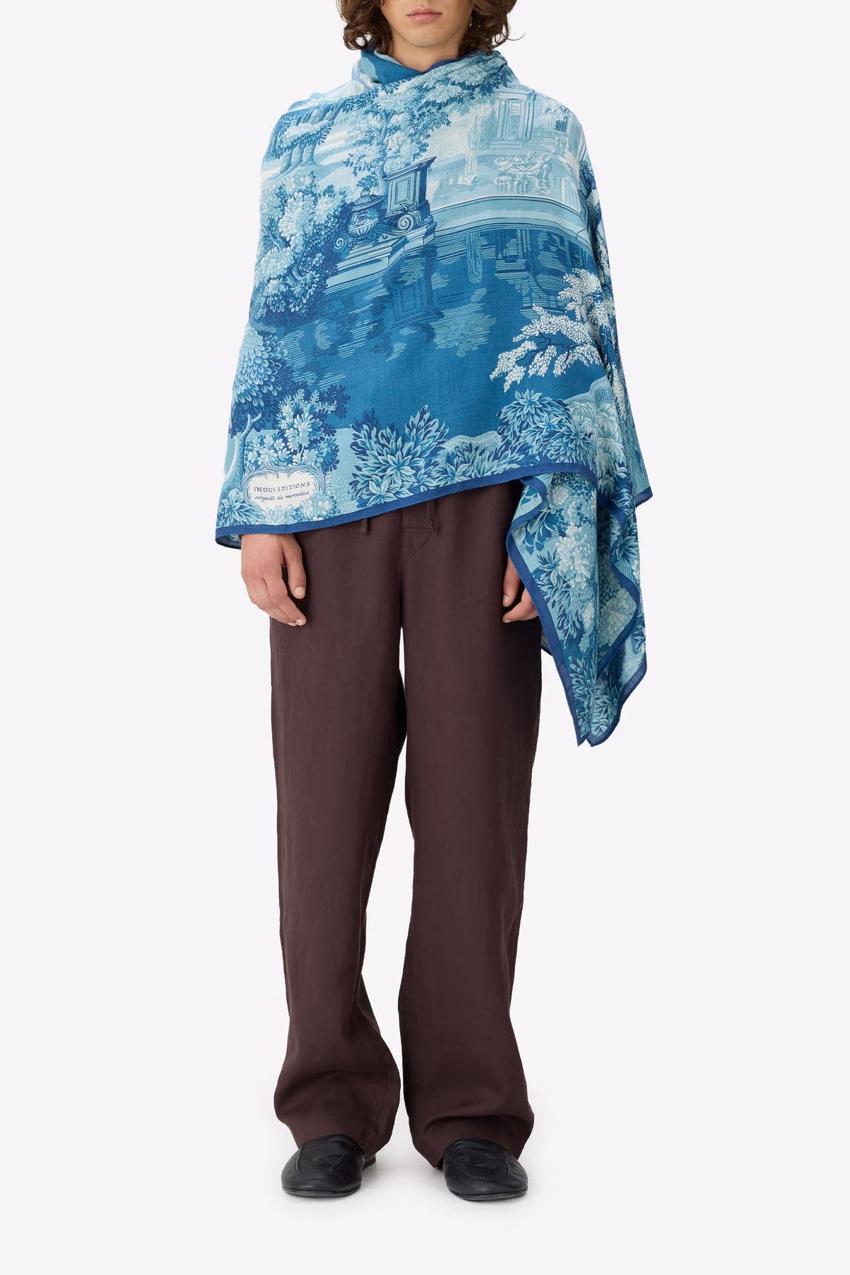A model stands against a light gray studio background wearing a blue and white toile-patterned poncho draped over their shoulders, paired with dark brown trousers and black shoes.