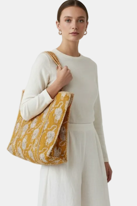 A woman wearing a cream knit long-sleeve top and matching skirt carries a large mustard yellow and white patterned woven tote bag against a plain light gray studio background.