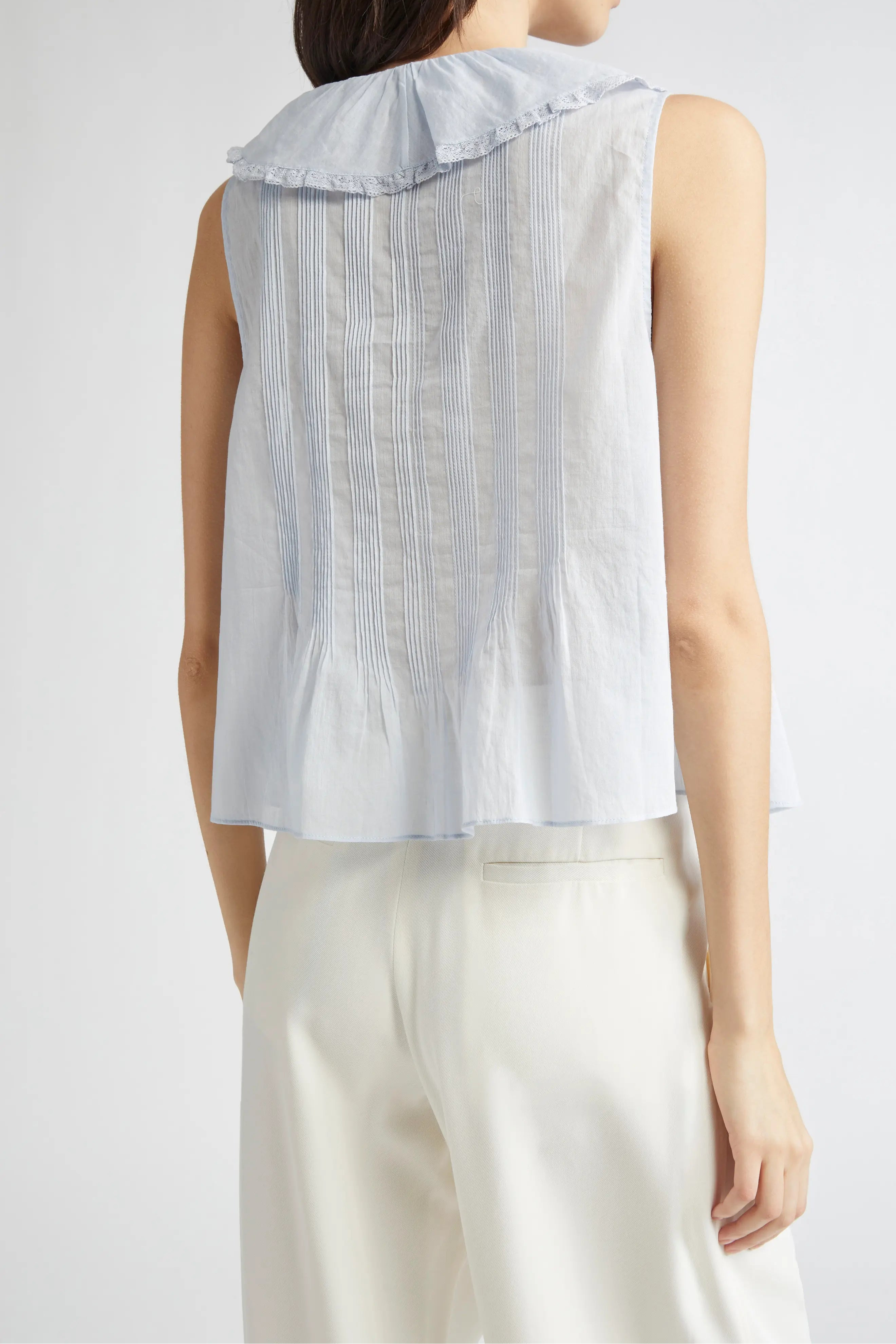 A sleeveless light blue top with a wide ruffled collar and vertical gathered detailing, shown from the back on a model wearing white pants against a light gray studio background.