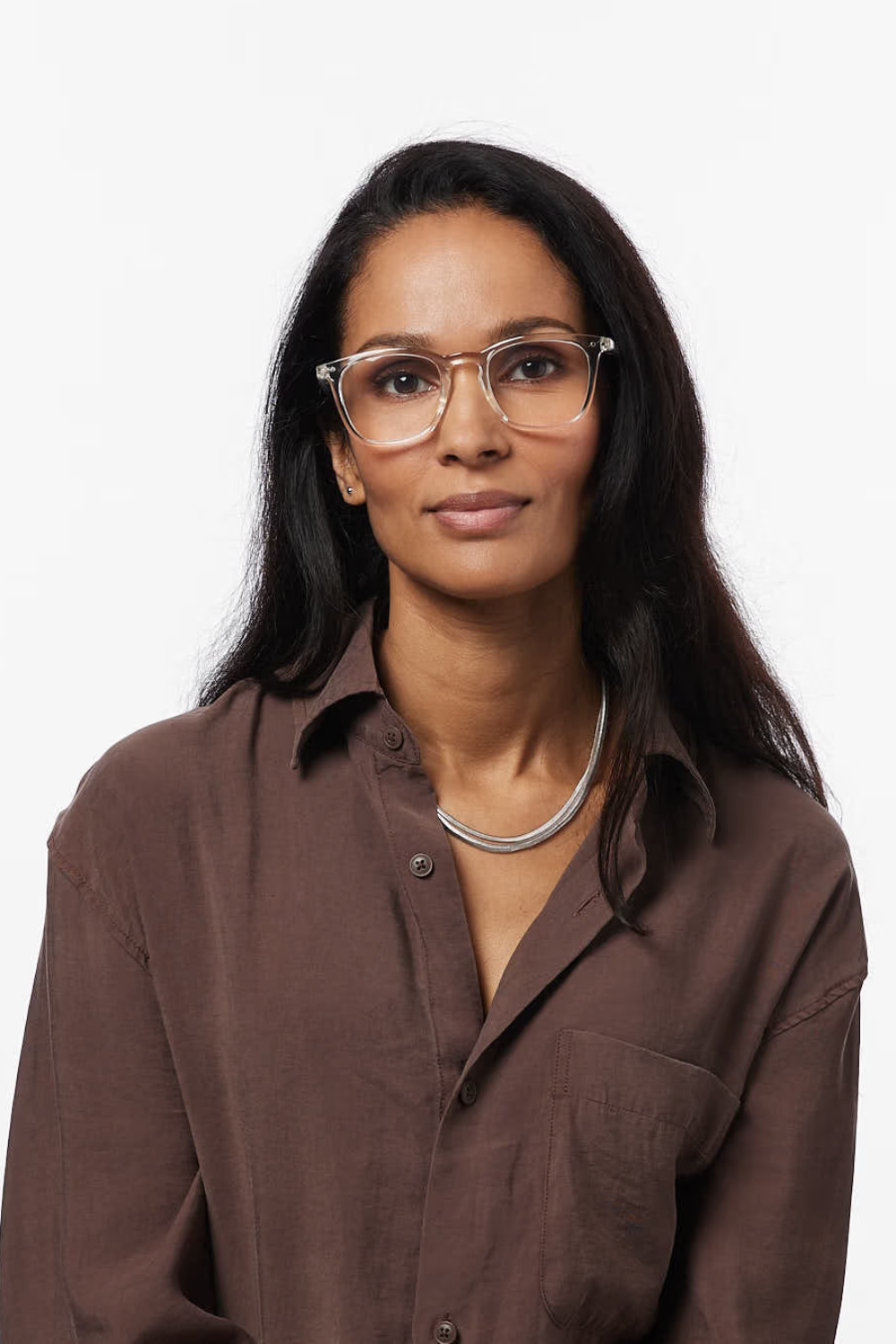 A woman with long dark hair wearing clear-framed eyeglasses and a brown button-up shirt, photographed against a plain light gray background.